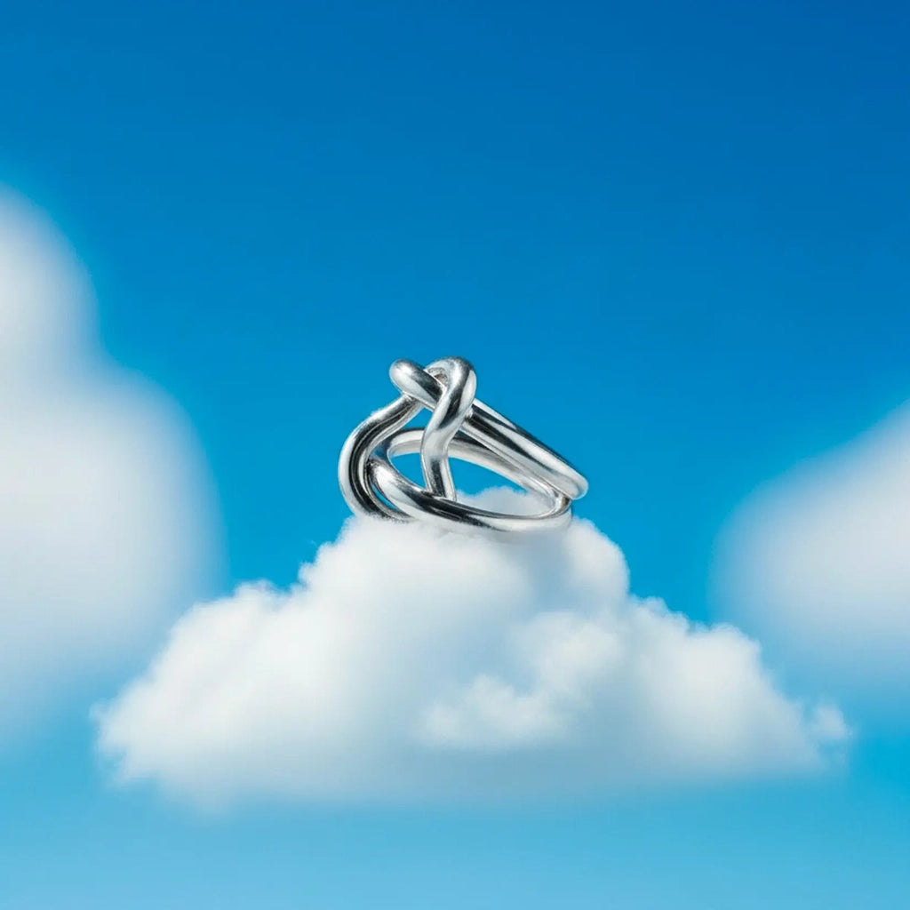 A silver ring with an interlocking knot design, resting on a fluffy white cloud against a bright blue sky background.